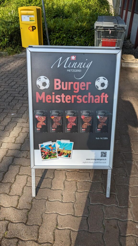 A sign in front of a shop reads "Burger Meisterschaft" which means tournament of burgers. It shows several burgers flavored according to countries playing at the Euro 2024