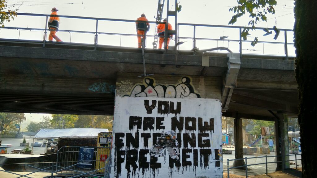 On a train overpass three workers in orange overalls, two seem to relax while one walks away. Below a sign reads: You Are Now Entering Free Reit