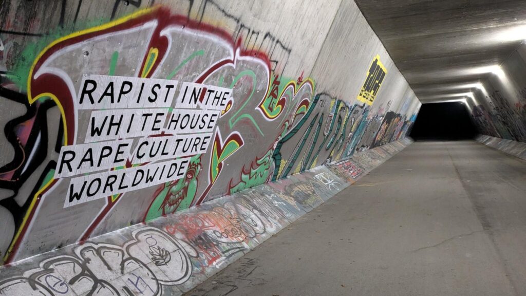 A tunnel is covered in graffitis, in the front a feminist collage reads: Rapist in the White House, Rape culture world wide