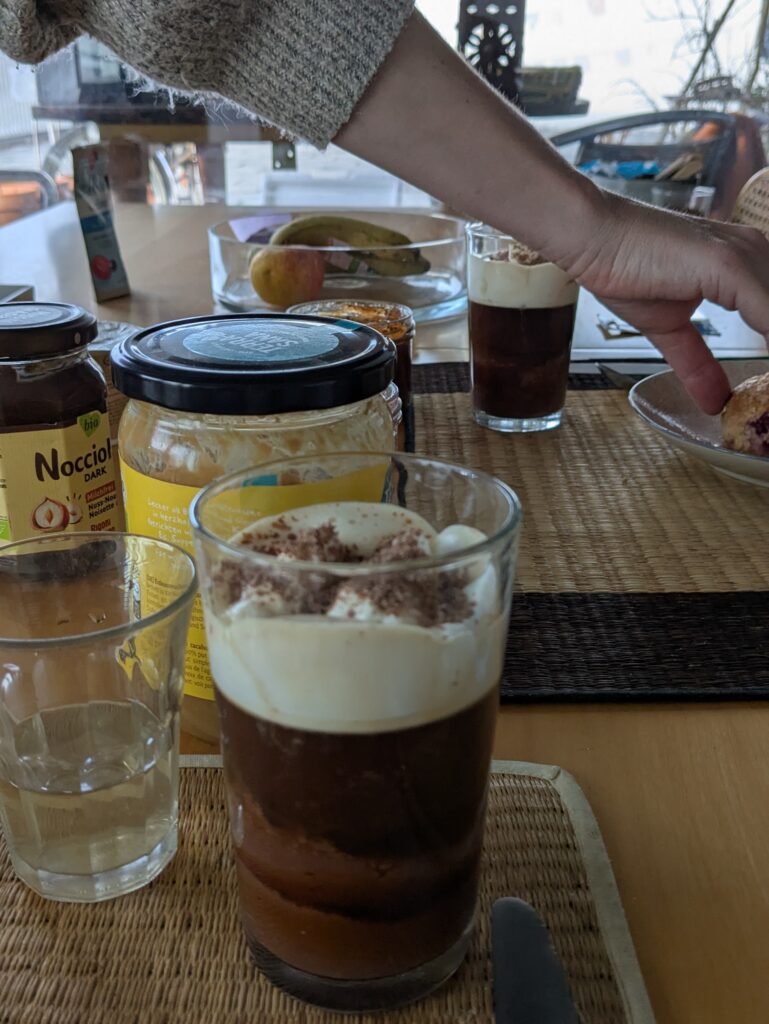 A breakfast table has a chocolate coffee drink with cream on top in front, in the back ground there is peanut butter and jams, a hand reaches across the table
