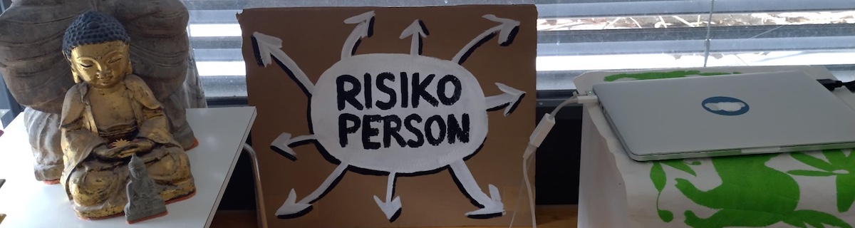 A sign reads "Risiko Person", which means person at risk, surrounded by arrows pointing in all directions. Some buddha statues and a laptop are visible