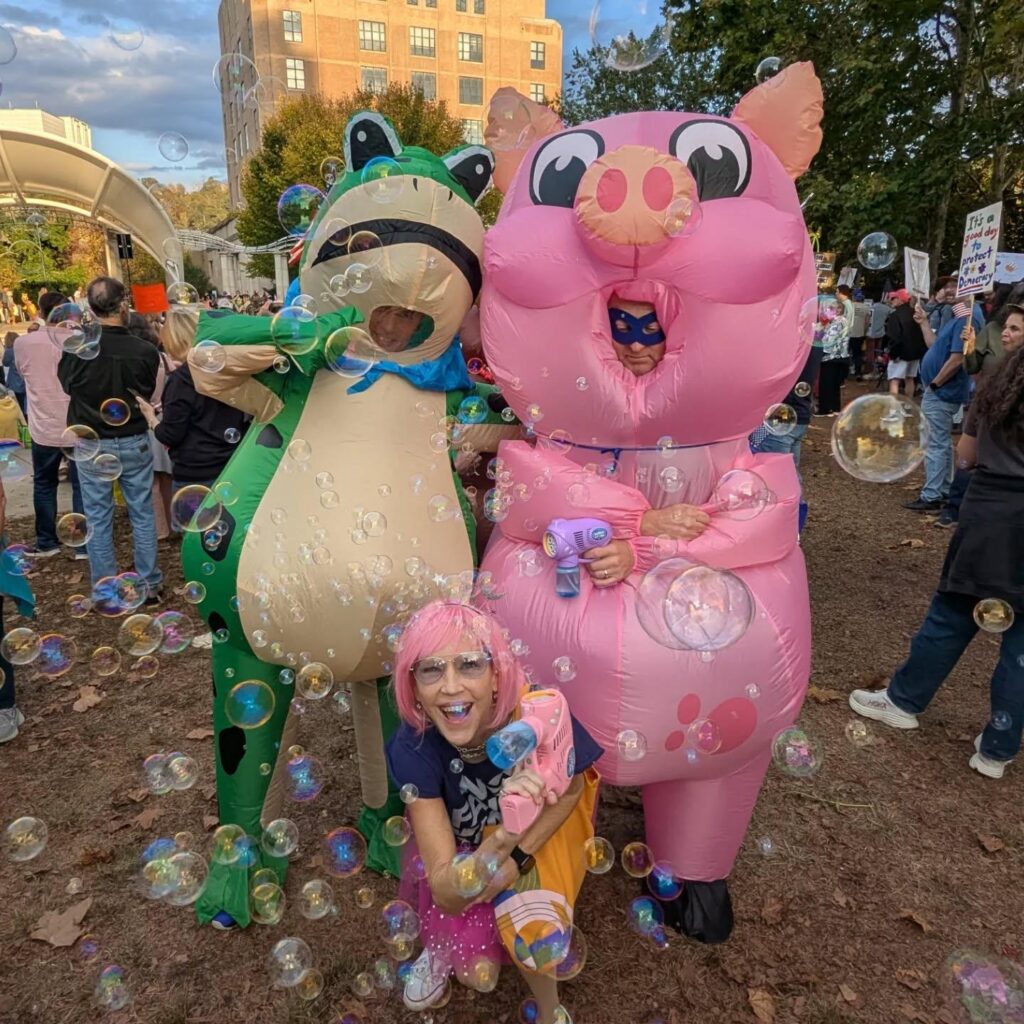A frog and a pig stand, a person with pink hair kneels in front of them, bubbles cover them