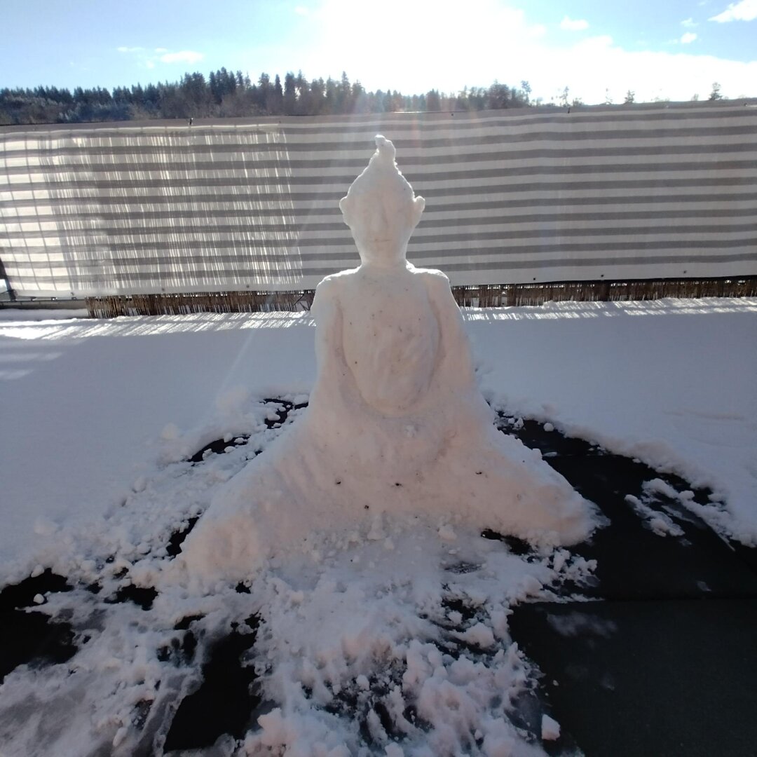 A Buddha figure made of snow sits on a sad balcony. In the background a striped tarp, a bit of forest and sunny sky