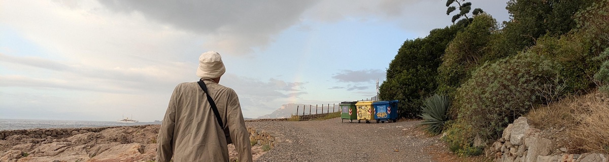 An old guy (me) walks away from an ocean beach, heading towards a cloudy sky and some trash cans